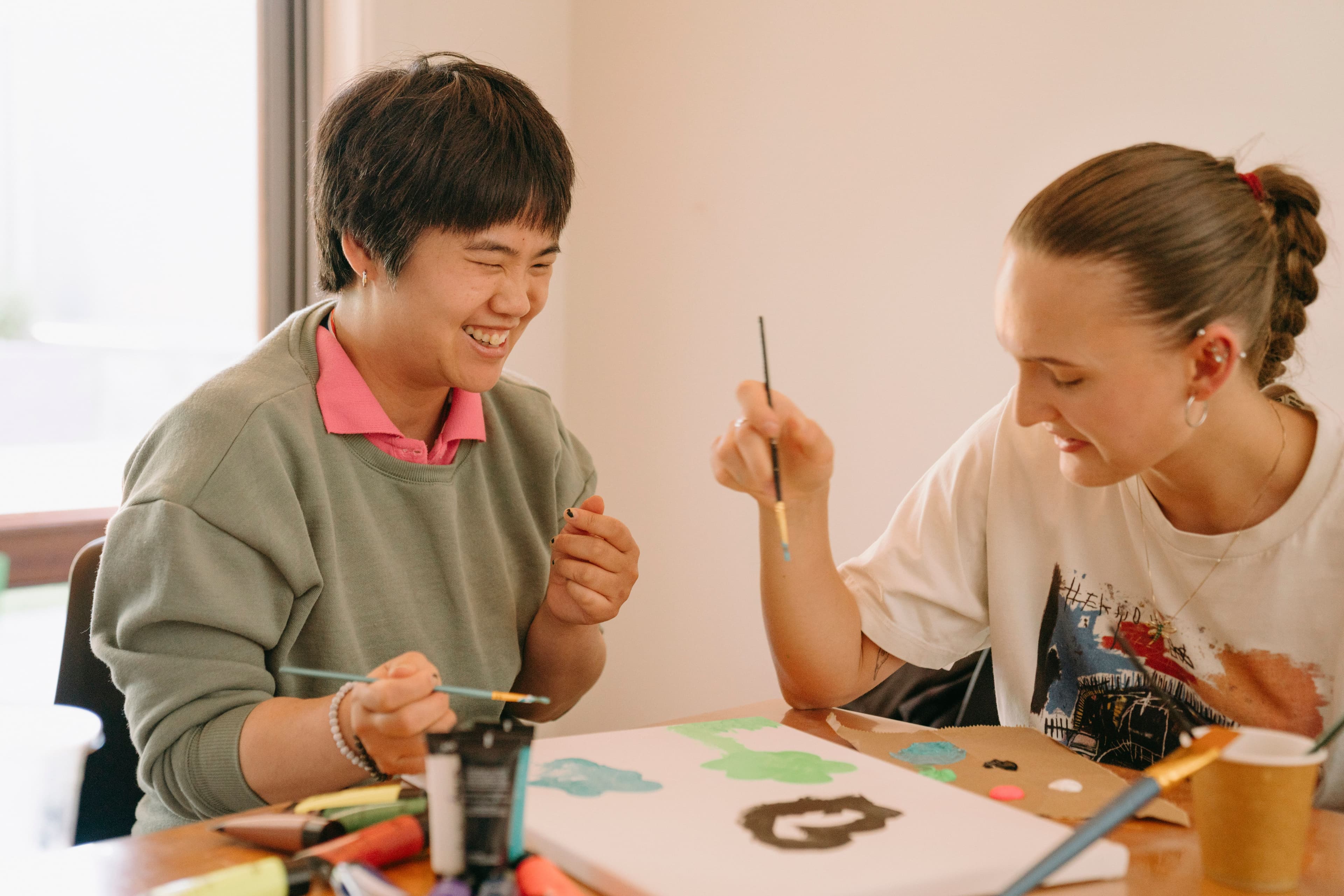 Melissa, who is an NDIS client, is smiling, holding a paintbrush and enjoying painting with a Bopha Cares disability worker who is also smiling and holding a paintbrush.